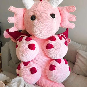 May include: A large, handmade pink crochet axolotl plush toy with white horns and red accents. The plush has large black eyes and is sitting on a grey sofa. The axolotl's body features red spots.