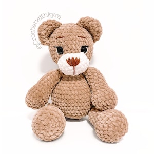 May include: A brown crocheted teddy bear with white and brown embroidered facial features. The bear is sitting with its legs outstretched.