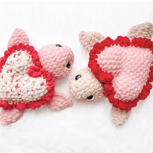 Op de afbeelding: Twee gebreide roze en rode hartvormige schildpad knuffels. De schildpadden hebben rode gebreide harten met witte accenten en roze gebreide schilden.