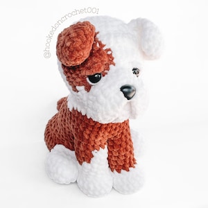 May include: A handmade crochet dog toy in a sitting pose. The toy is predominantly brown and white, with brown detailing on the ears and back. It has black eyes and a black nose. The text "@hookedoncrochet001" is visible on the image.