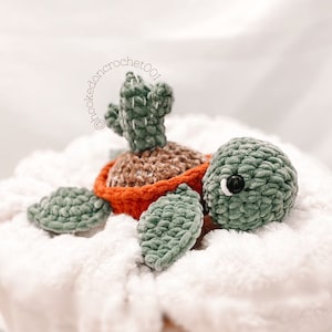 May include: A handmade crochet turtle with a cactus on its back, resting on a fluffy white surface. The turtle is green with a black eye, and the cactus sits in an orange pot. The turtle's flippers are also green. The base is a woven basket.