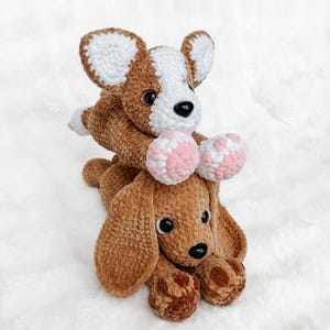 May include: A brown and white crocheted corgi plush toy resting on top of a brown crocheted basset hound plush toy. Both stuffed animals feature dark plastic eyes, black noses, and detailed paw pads in pink and brown yarn textures.