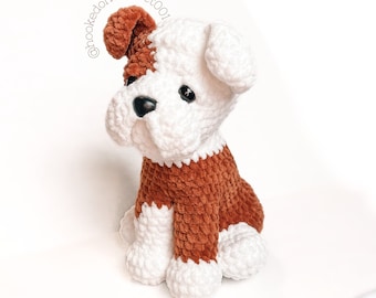 May include: A plush crochet dog toy featuring a white and brown color pattern, black plastic eyes, and a black nose. The dog is in a sitting position with one ear folded down. The text @hookedoncrochet001 is visible above the dog's head.
