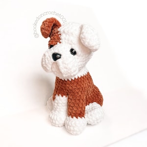 May include: A handmade crochet dog plushie in brown and white. The dog has a brown body and head patch, with white accents on the face, paws, and ears. Black safety eyes and a white collar complete the design. The text "@hookedoncrochet001" is in the upper left corner.