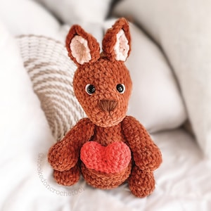 May include: A brown crocheted kangaroo holding a red heart. The kangaroo is sitting on a white surface. The text "@hooked.on.crochet001" is written on the surface.