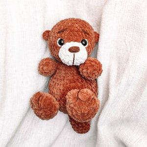 May include: A brown crocheted teddy bear with white facial features and black eyes. The bear is lying on a white fuzzy blanket.