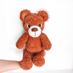 Peut inclure: Un ours en peluche crochet&eacute; marron avec des accents blancs autour des yeux et du nez. L'ours est assis sur une main.