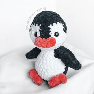 May include: A crocheted penguin toy with black and white fur, red beak and feet. The penguin is sitting on a white surface.