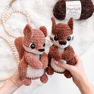 Könnte beinhalten: Zwei handgefertigte gehäkelte Eichhörnchen-Plüschtiere in Braun- und Cremetönen. Die Eichhörnchen haben schwarze Augen und werden vor einem weißen, flauschigen Hintergrund gehalten. Zwei Knäuel braunes Garn sind im Hintergrund zu sehen, mit dem Text "Premier Parfait Chunky".