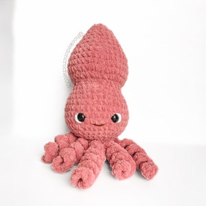 Puede incluir: Un juguete de peluche de calamar tejido a crochet de color rosa con ojos negros y una boca sonriente. El calamar tiene ocho tentáculos.