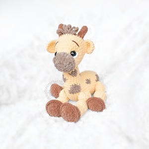May include: A crocheted giraffe toy with brown spots and a light brown body. The giraffe has brown fur on its legs and face. The toy is sitting on a white fluffy surface.