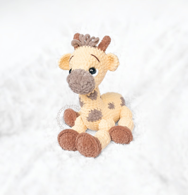 May include: A crocheted giraffe toy with brown spots and a light brown body. The giraffe has brown fur on its legs and face. The toy is sitting on a white fluffy surface.