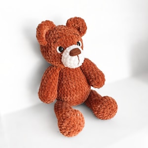Peut inclure: Un ours en peluche crochet&eacute; marron avec un visage blanc et des yeux noirs. L'ours est assis sur une surface blanche.