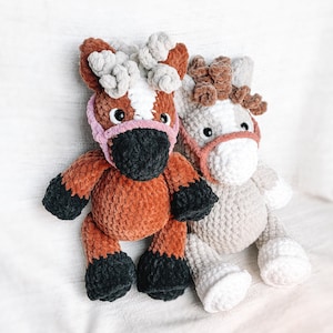 May include: Two crocheted plush horse toys. One is brown with a white stripe and a pink nose, the other is beige with a brown mane and a pink nose.