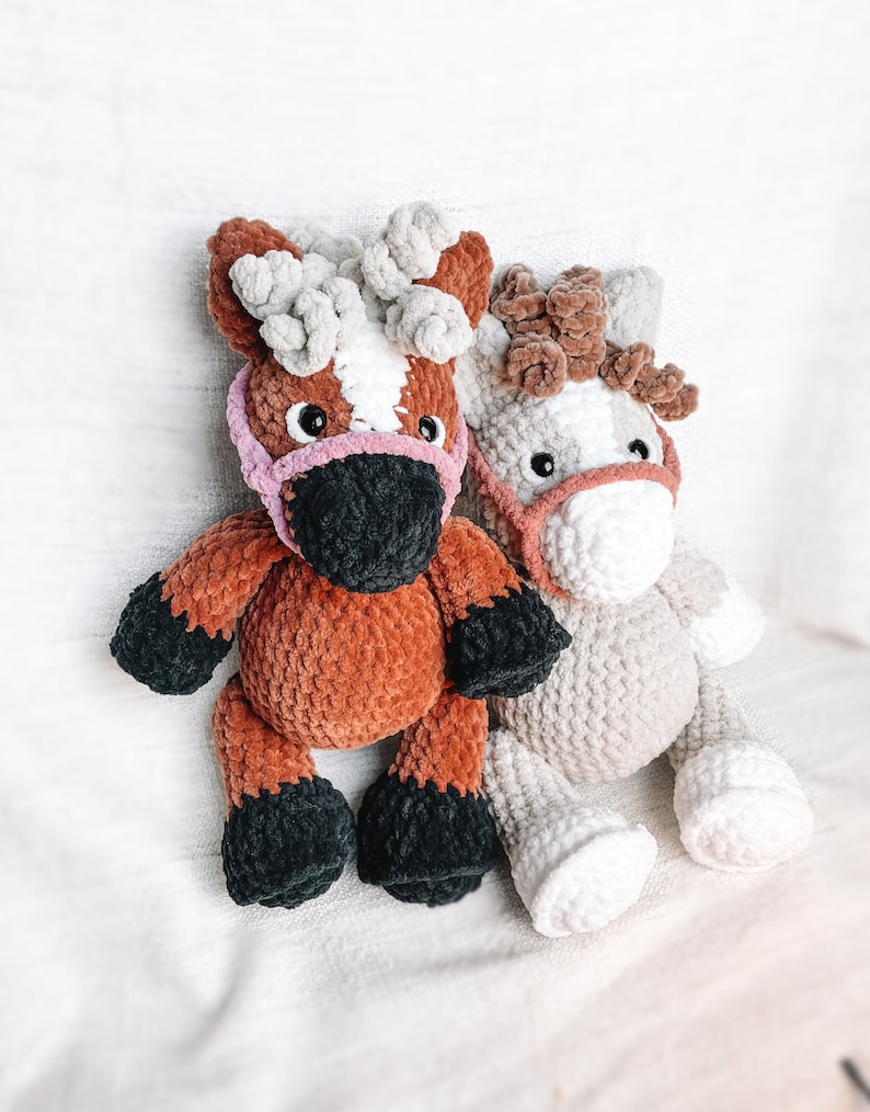 May include: Two crocheted plush horse toys. One is brown with a white stripe and a pink nose, the other is beige with a brown mane and a pink nose.