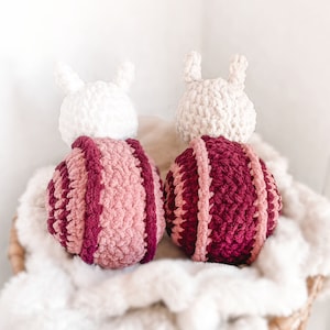 K&ouml;nnte beinhalten: Zwei geh&auml;kelte Stoffschnecken mit wei&szlig;en K&ouml;pfen und rosa-burgunderfarbenen gestreiften K&ouml;rpern. Die Schnecken sitzen auf einer wei&szlig;en, flauschigen Decke.