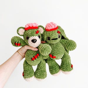 Puede incluir: Dos peluches, un oso y un conejo, hechos de hilo texturizado verde. Ambos presentan partes superiores rosas similares a cerebros, ojos de seguridad negros y parches cosidos rojos en extremidades y cabeza. El oso tiene hocico y patas crema.