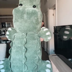 May include: A large, soft, green crocheted alligator plush toy. The alligator has white accents on its feet and a textured, scaly design along its back. The toy is being held up by a person. The alligator's eyes are black.