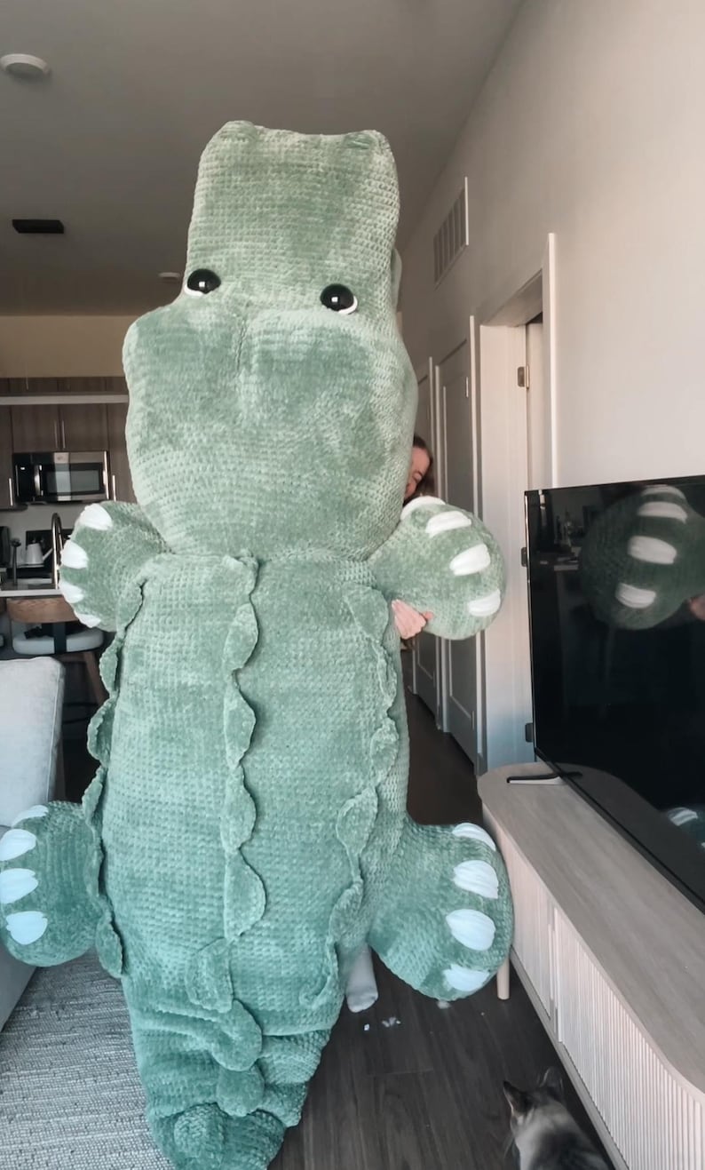 May include: A large, soft, green crocheted alligator plush toy. The alligator has white accents on its feet and a textured, scaly design along its back. The toy is being held up by a person. The alligator's eyes are black.