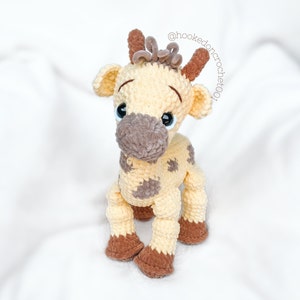 May include: A crocheted giraffe toy with brown and beige yarn. The giraffe has large brown eyes and a brown nose. The giraffe is sitting on a white surface.