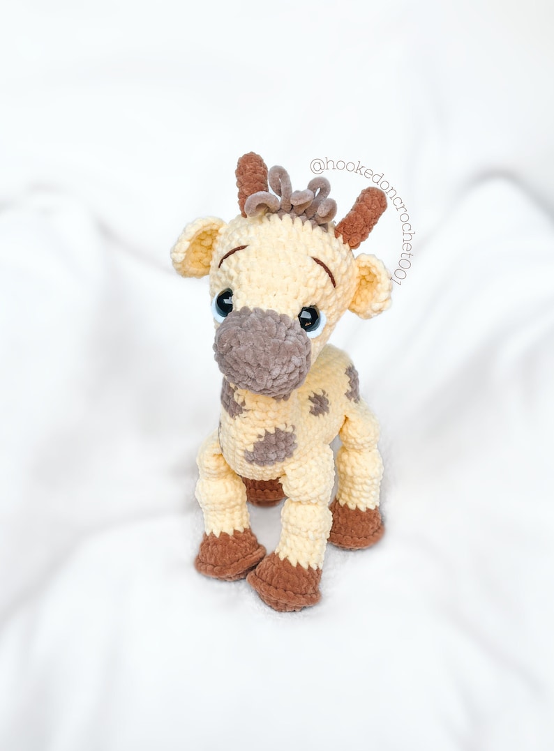 May include: A crocheted giraffe toy with brown and beige yarn. The giraffe has large brown eyes and a brown nose. The giraffe is sitting on a white surface.