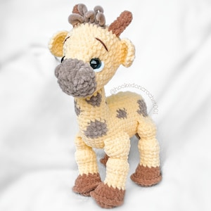 May include: A crocheted giraffe toy with brown and yellow yarn. The giraffe has brown spots and a brown nose. The giraffe's eyes are blue and the toy is standing on a white surface. The text "@hookedoncrochet001" is visible on the toy.