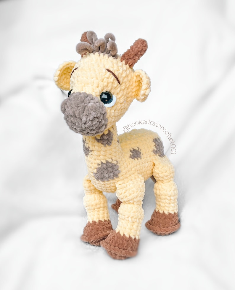 May include: A crocheted giraffe toy with brown and yellow yarn. The giraffe has brown spots and a brown nose. The giraffe's eyes are blue and the toy is standing on a white surface. The text "@hookedoncrochet001" is visible on the toy.