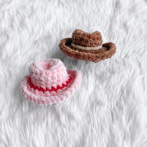 May include: Two miniature crochet cowboy hats. One is light pink with a red brim, and the other is brown with a tan band. The hats are made of a soft, textured yarn and are likely intended for a baby or small pet.