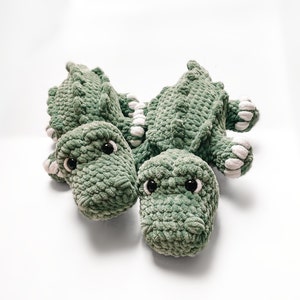 May include: Two green crocheted alligator plushies with white accents and black eyes. The plushies are lying on a white surface.