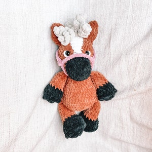 May include: A crocheted brown and black horse plush toy with a pink nose and white mane. The horse is wearing a pink crocheted bridle.