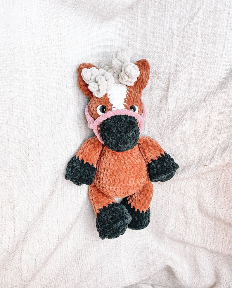 May include: A crocheted brown and black horse plush toy with a pink nose and white mane. The horse is wearing a pink crocheted bridle.