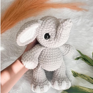May include: A handmade gray crochet elephant plush toy with large ears and black eyes. The elephant is soft and cuddly, perfect for a child's room or nursery. The plush toy is made with a textured yarn.
