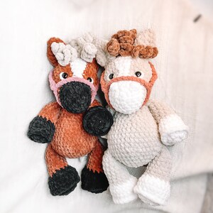 May include: Two crocheted stuffed horses, one brown and one beige, with pink noses and black eyes. The horses are sitting on a white fuzzy surface.
