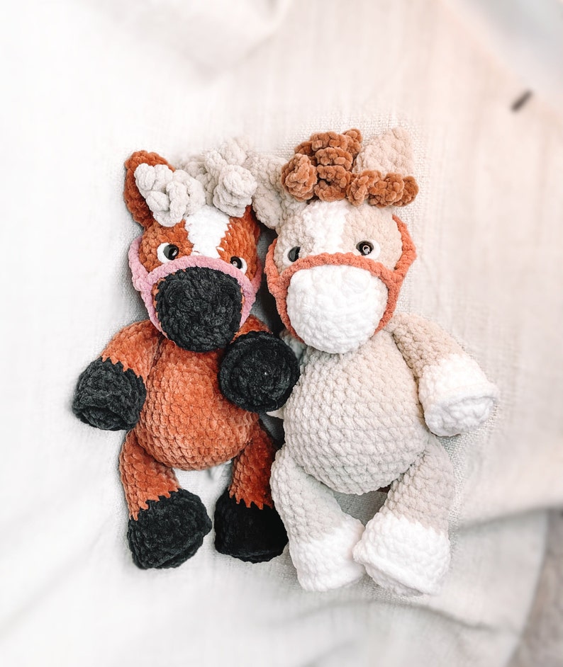 May include: Two crocheted stuffed horses, one brown and one beige, with pink noses and black eyes. The horses are sitting on a white fuzzy surface.