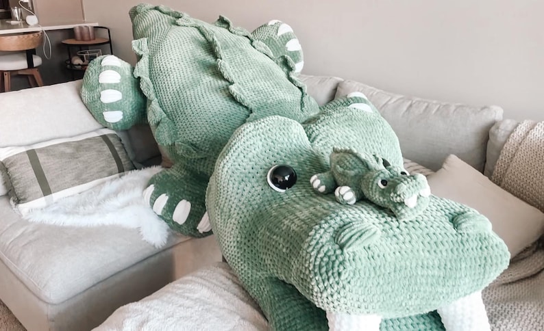 May include: A large, soft, green alligator plush toy with white accents on its feet and a smaller alligator plush. The toys are on a light gray couch with pillows and a white faux fur throw. The alligator has a textured, knitted appearance.