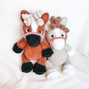 May include: Two crocheted horse toys. One is brown with black hooves and a pink nose. The other is gray with white hooves and a pink nose.