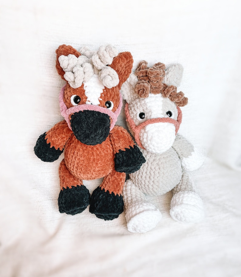 May include: Two crocheted horse toys. One is brown with black hooves and a pink nose. The other is gray with white hooves and a pink nose.