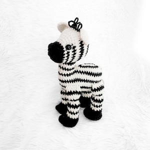 May include: A crocheted zebra stuffed animal with black and white stripes. The zebra has a black nose and black eyes. The zebra is standing on a white fluffy surface.
