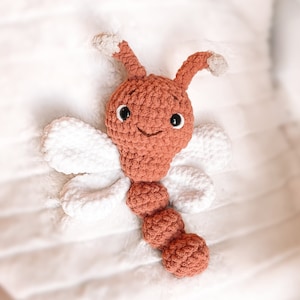 May include: A crocheted dragonfly toy featuring a rust-colored head and segmented body, two antennae with light beige tips, and four white wings. The toy has black plastic safety eyes and a stitched mouth, crafted from soft yarn.