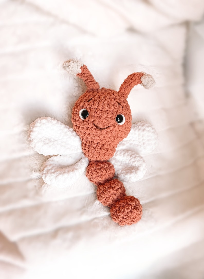 May include: A crocheted dragonfly toy featuring a rust-colored head and segmented body, two antennae with light beige tips, and four white wings. The toy has black plastic safety eyes and a stitched mouth, crafted from soft yarn.