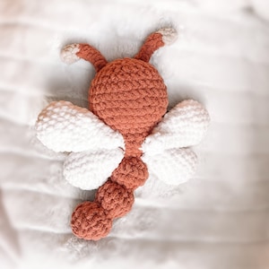 May include: A crocheted dragonfly toy featuring a rust-colored head, body, and tail with two pairs of white wings. The head includes two rust-colored antennae with light beige tips. The body is segmented into three rounded sections.
