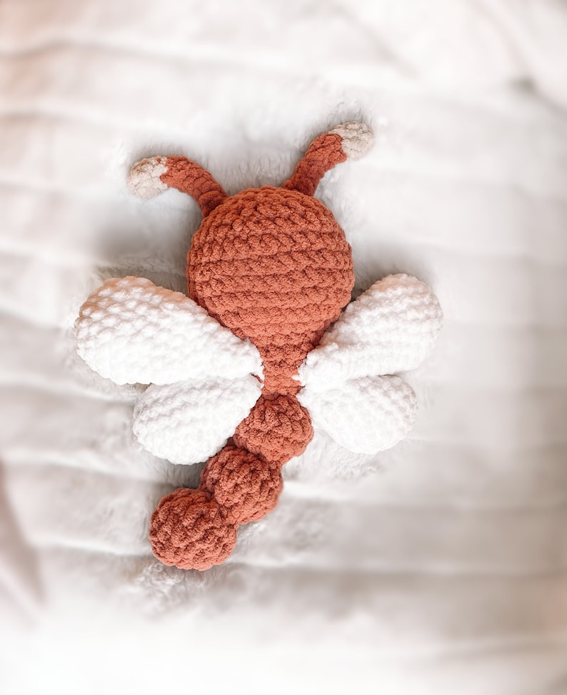 May include: A crocheted dragonfly toy featuring a rust-colored head, body, and tail with two pairs of white wings. The head includes two rust-colored antennae with light beige tips. The body is segmented into three rounded sections.