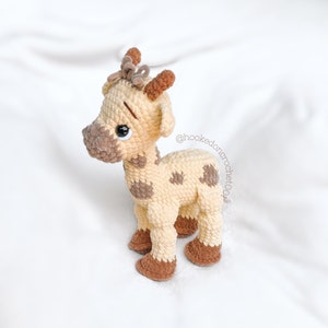 May include: A crocheted giraffe toy with brown spots and a brown bow on its head. The giraffe is standing on a white surface. @hooked.on.crochet