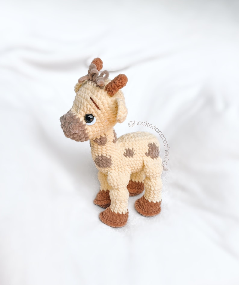 May include: A crocheted giraffe toy with brown spots and a brown bow on its head. The giraffe is standing on a white surface. @hooked.on.crochet