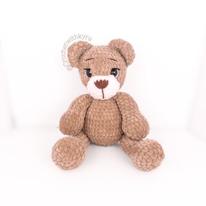 Peut inclure: Un ours en peluche crochet&eacute; marron avec des traits du visage brod&eacute;s en blanc et marron. L'ours est assis les jambes crois&eacute;es.