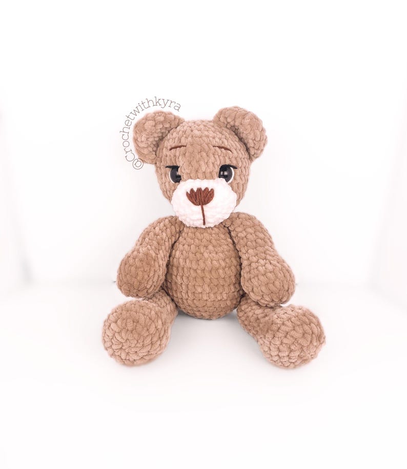 Peut inclure: Un ours en peluche crochet&eacute; marron avec des traits du visage brod&eacute;s en blanc et marron. L'ours est assis les jambes crois&eacute;es.
