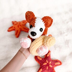 Puede incluir: Un perro de peluche tejido a ganchillo con hilo naranja óxido y blanco, con ojos de seguridad negros y nariz negra. El perro sostiene un anillo tejido a ganchillo de color amarillo claro. Dos estrellas de mar tejidas en tonos naranja y rojo.