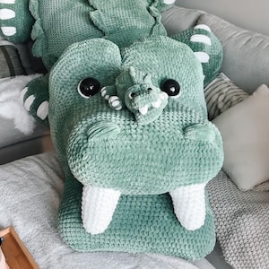May include: A large, plush, green crocodile-shaped pillow with white teeth and black eyes. The pillow is on a gray couch with other pillows and blankets. The crocodile has a smaller crocodile on its snout.