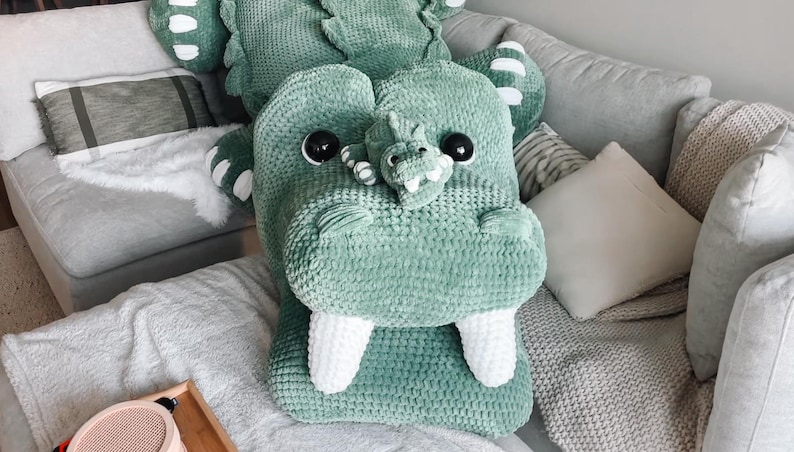 May include: A large, plush, green crocodile-shaped pillow with white teeth and black eyes. The pillow is on a gray couch with other pillows and blankets. The crocodile has a smaller crocodile on its snout.