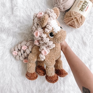 May include: A handmade crochet horse plushie in shades of brown, tan, and cream, with pink accents. The plush toy features a fluffy mane and tail, and is made with soft, textured yarn. The horse is a cuddly toy.
